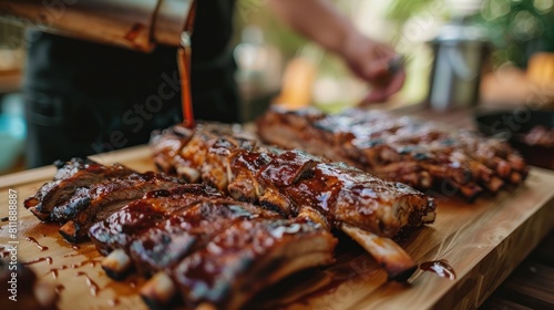 Grilled ribs are laid on a wooden cutting board, and in the backdrop is a blurry high angle shot of an unnamed cook pouring barbecue photo