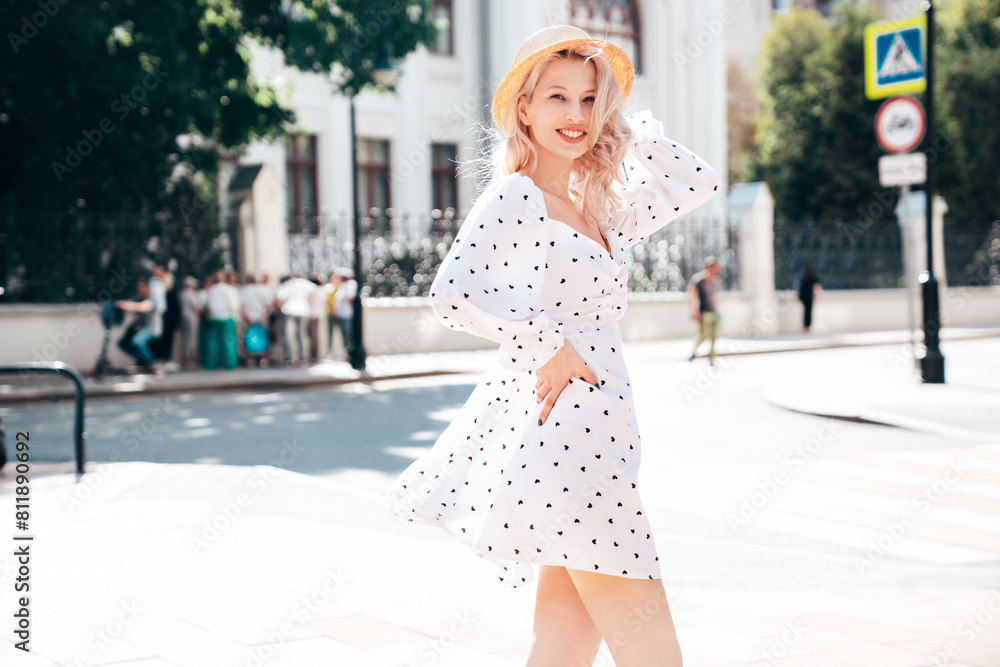 Naklejka premium Young beautiful smiling blond woman in trendy summer white dress. Sexy carefree woman posing in street at sunset. Positive model outdoors at sunny day. Cheerful and happy. In hat