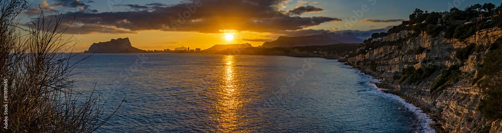 Golden sunset paints the sky as it dips below the mountainous horizon, reflecting on the Mediterranean waters near the coastal cliffs of Calpe, Spain Golden sunset paints the sky as it dips below the mountainous horizon, reflecting on the Mediterranean waters near the coastal cliffs of Calpe, Spain