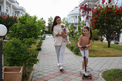 Happy mom and daughter walking their rough coated jack russell terrier. Little girl riding a push scooter with her mother and wire haired pup. Copy space, background.