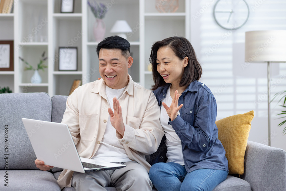 Cheerful Asian couple enjoying a lively video call from their cozy living room. They sit side by side on a grey sofa, laughing and waving at the laptop screen, exuding happiness and connection Cheerful Asian couple enjoying a lively video call from their cozy living room. They sit side by side on a grey sofa, laughing and waving at the laptop screen, exuding happiness and connection
