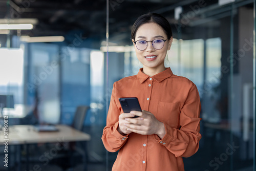 Confident Asian businesswoman in stylish orange blouse using a smartphone in a well-lit modern office environment. Perfect for professional and technology themes.