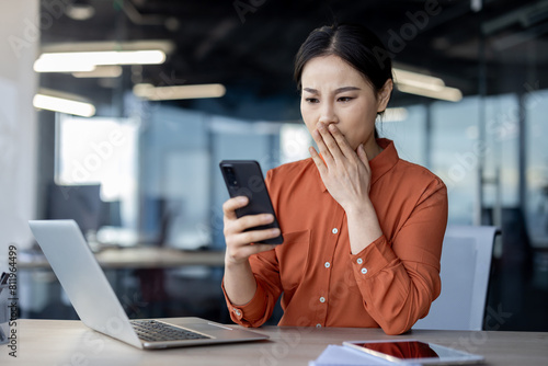 Asian woman in orange blouse looks dismayed at phone in modern office setting. Reacting to surprising bad news. photo