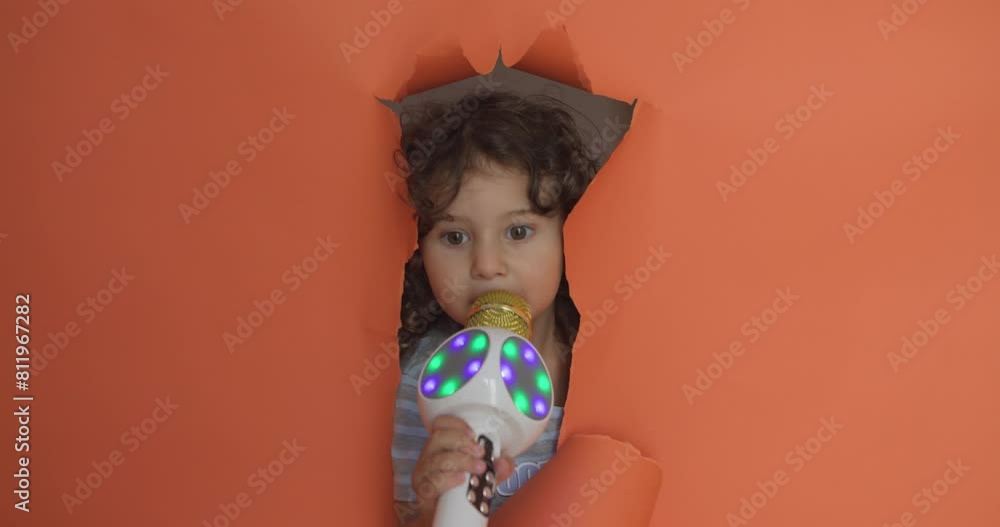 A playful boy pops his face through a torn orange paper backdrop with a bright LED-lit microphone in hand, suggesting a fun karaoke or singing moment. A playful boy pops his face through a torn orange paper backdrop with a bright LED-lit microphone in hand, suggesting a fun karaoke or singing moment.