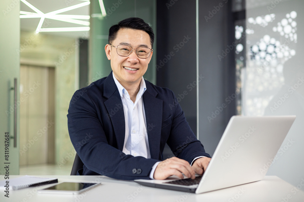 A professional Asian businessman smiles as he engages with work on his laptop in a sleek, well-lit office environment, portraying efficiency and positivity. A professional Asian businessman smiles as he engages with work on his laptop in a sleek, well-lit office environment, portraying efficiency and positivity.