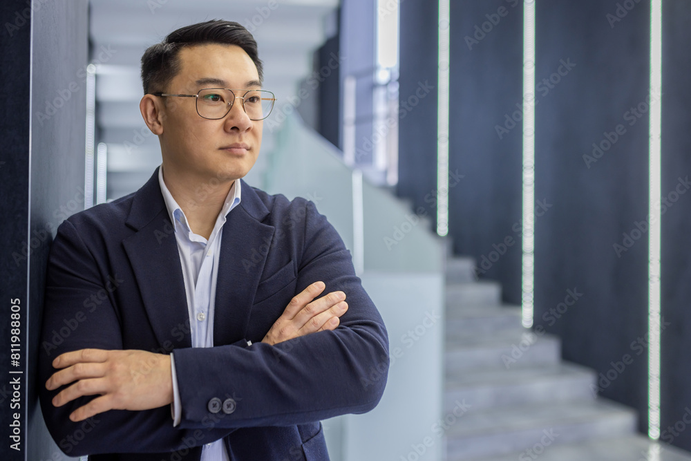 A thoughtful and confident Asian businessman with arms crossed, standing by a staircase in a sleek, modern office environment. A thoughtful and confident Asian businessman with arms crossed, standing by a staircase in a sleek, modern office environment.