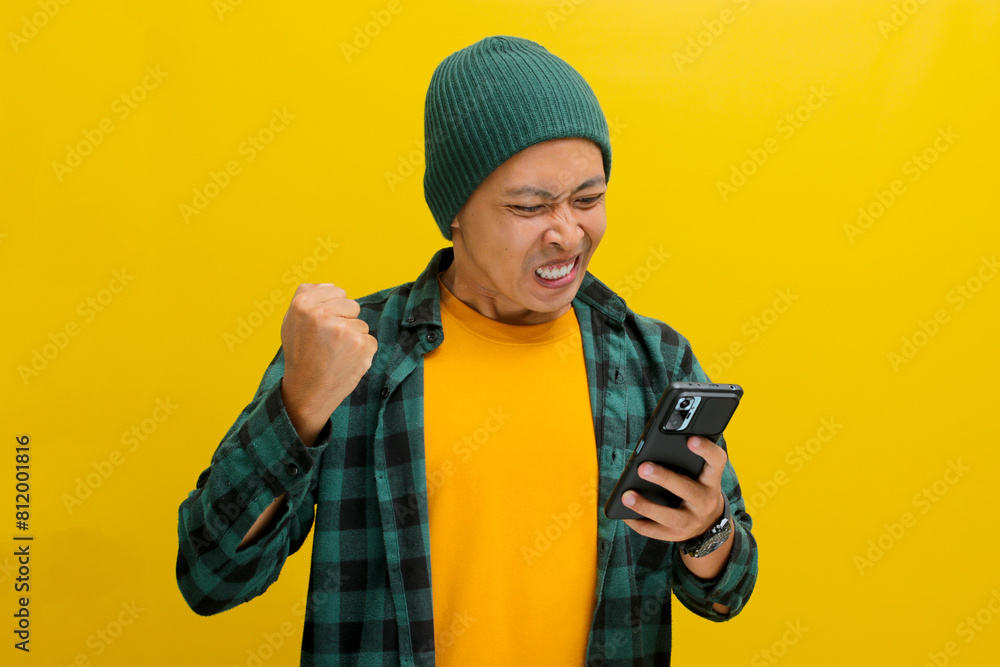 An angry Asian man, dressed in a beanie hat and casual shirt, visibly reacts to bad news while looking at his mobile phone, while standing against yellow background An angry Asian man, dressed in a beanie hat and casual shirt, visibly reacts to bad news while looking at his mobile phone, while standing against yellow background