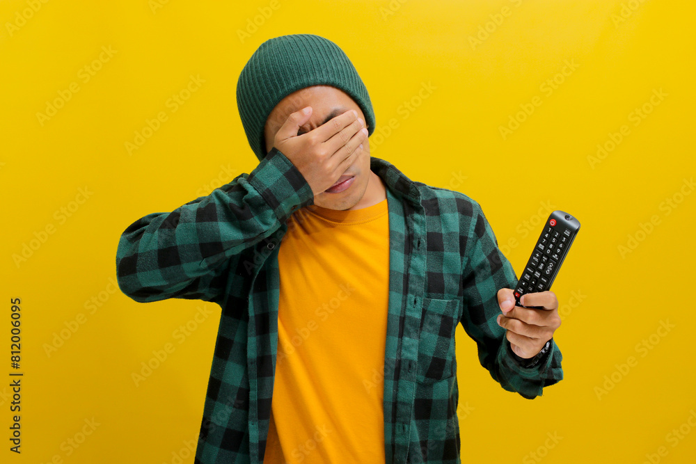 Anxious young Asian man, dressed in a beanie hat and casual shirt, appears stressed while holding a TV remote, his face resting in his palm, while standing against yellow background Anxious young Asian man, dressed in a beanie hat and casual shirt, appears stressed while holding a TV remote, his face resting in his palm, while standing against yellow background
