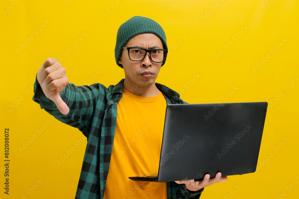A displeased young Asian man, dressed in a beanie hat and casual shirt, expresses dissatisfaction with a thumbs down gesture while holding a laptop in his hand, isolated on a yellow background A displeased young Asian man, dressed in a beanie hat and casual shirt, expresses dissatisfaction with a thumbs down gesture while holding a laptop in his hand, isolated on a yellow background