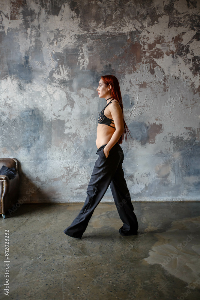 A beautiful red-haired girl in sexy lingerie and black pants poses in a concrete room.