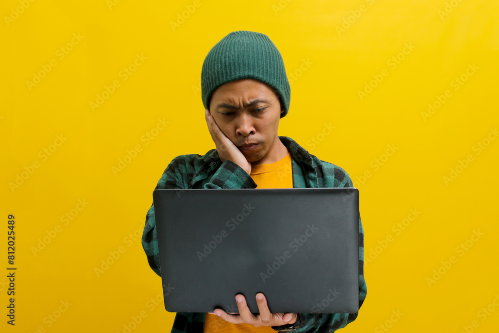 A concerned young Asian male student or worker appears bored and worried while looking at a laptop computer, watching an online training course, Isolated on a yellow background. A concerned young Asian male student or worker appears bored and worried while looking at a laptop computer, watching an online training course, Isolated on a yellow background.