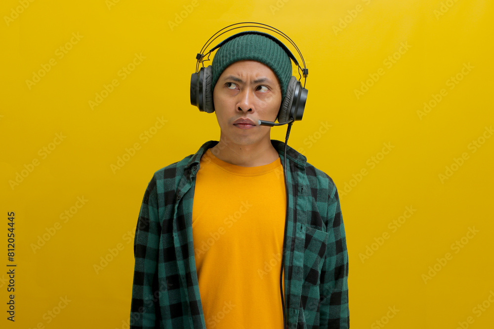 Asian man in a casual clothes, wearing headphones, looks up in deep thought, seemingly troubled by something on his music. A worried expression creases his brow. Isolated on a yellow background. Asian man in a casual clothes, wearing headphones, looks up in deep thought, seemingly troubled by something on his music. A worried expression creases his brow. Isolated on a yellow background.