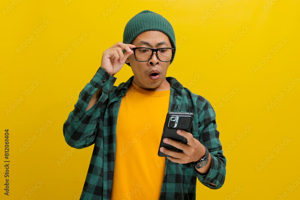 Shocked Asian man, sporting a beanie hat and casual shirt, adjusts his glasses while gazing at his smartphone with a surprised expression, exclaiming Wow, Great News Standing against yellow background Shocked Asian man, sporting a beanie hat and casual shirt, adjusts his glasses while gazing at his smartphone with a surprised expression, exclaiming Wow, Great News Standing against yellow background
