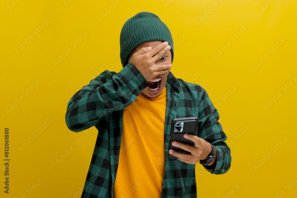 Shocked Asian man, dressed in a beanie hat and casual shirt, covers his eyes in a comical and surprised reaction while looking at his phone, clearly taken aback by unexpected news, yellow background Shocked Asian man, dressed in a beanie hat and casual shirt, covers his eyes in a comical and surprised reaction while looking at his phone, clearly taken aback by unexpected news, yellow background