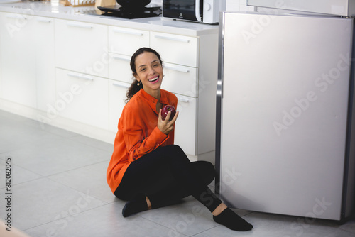 Beautiful African Latin American female is enjoy eating and holding a red apple from refrigerator, concept of freshness, healthy food, vegetable, nourishment at home, refreshment from fruit in fridge