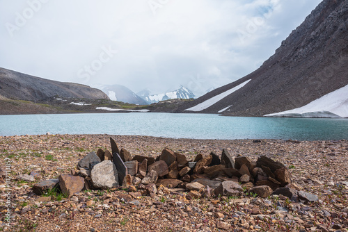 Sharp pointed stones on background azure alpine lake against snow-covered few pointy peaks in low clouds. Ripples on blue water surface of mountain lake and three snowy peaked tops in low cloudiness.