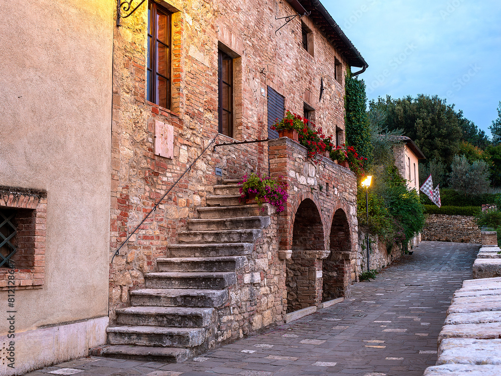 Medieval stone houses at dawn in an ancient thermal village in the Val d'Orcia in Tuscany, with lights still on, creating a warm fairytale atmosphere. From one house hang two plaid banners. Medieval stone houses at dawn in an ancient thermal village in the Val d'Orcia in Tuscany, with lights still on, creating a warm fairytale atmosphere. From one house hang two plaid banners.
