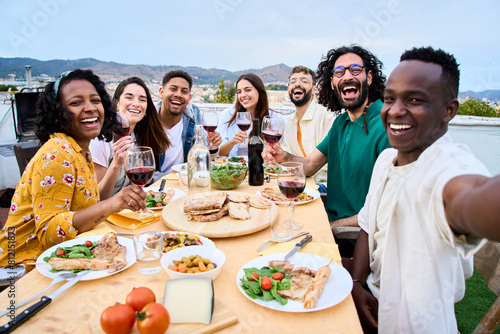 Young African smiling man taking selfie to joyful millennial people sitting at barbecue table. Portrait of summer rooftop meeting group of multiracial friends posing happy looking at camera outdoors 