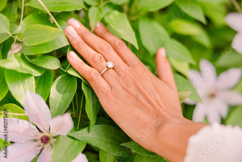 A tan skin woman has her hands over leaves and greenery in a garden. She wears a solitary diamond engagement ring.