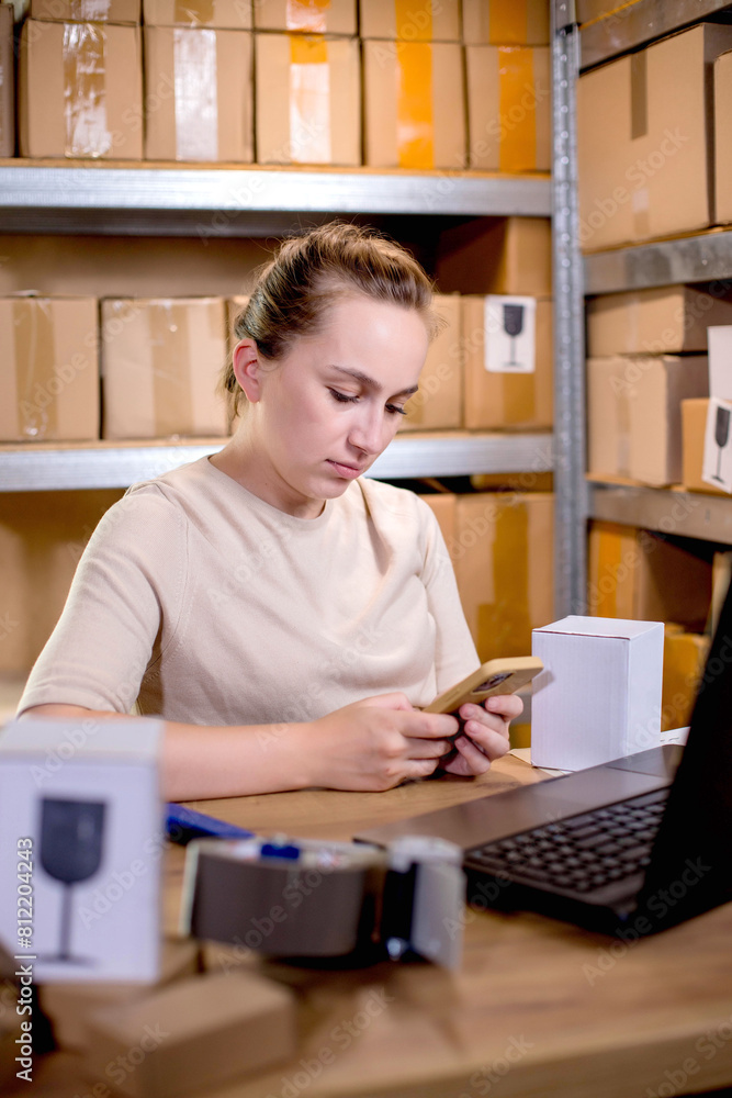 Fototapeta premium Young entrepreneur small business checking internet orders on smartphone and preparing boxed items for delivery to customers
