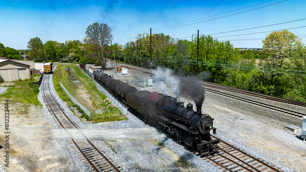 Obraz premium Aerial vintage steam train moves through industrial rail yard, emitting steam as it passes warehouses alongside other rail cars