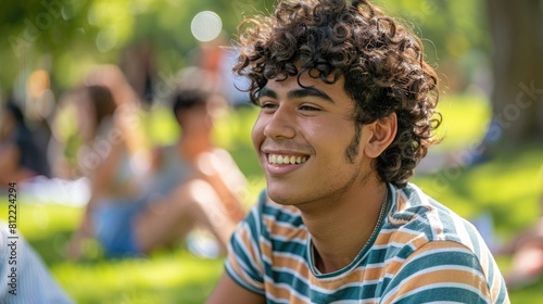A young man with curly hair donning a striped shirt flashes a bright smile as he sits in the park on a sunny summer day engrossed in conversation with friends and glued to his cellphone dur