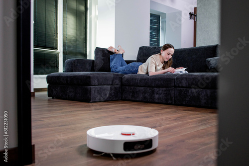 Woman lying on sofa and reading a book while robot vacuum cleaner doing housework