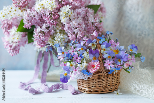 Spring still life with flowers, a bouquet of blooming lilacs and bird cherry in a vase, a basket with pansies, forget-me-nots and phlox, a beautiful postcard.