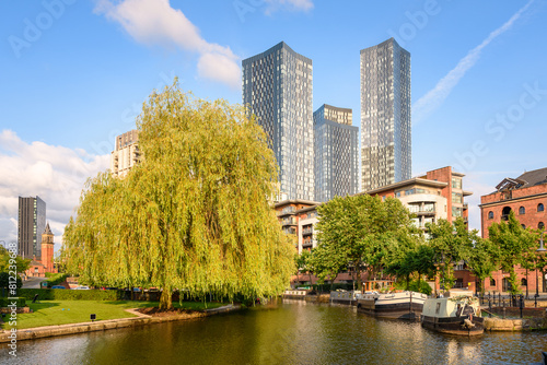 Modern glass residential towers and apartment blocks overlooking a canal with moored barges on a clear summer day. 