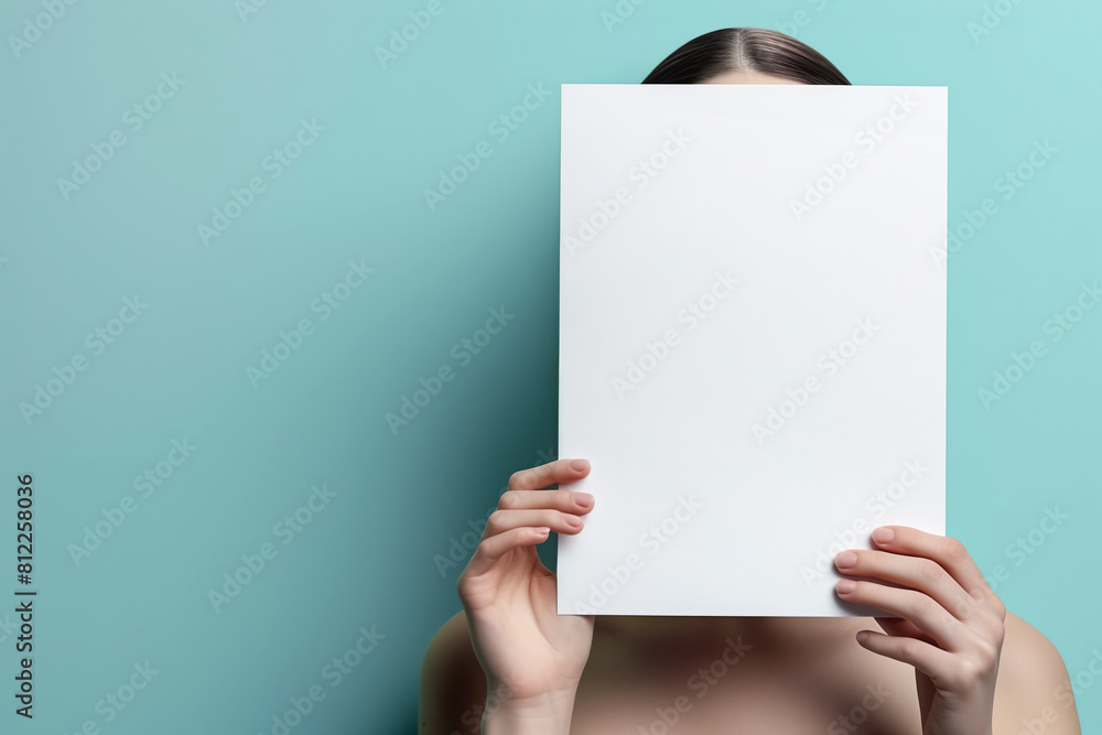 young female model holding blank sheet white empty paper in front of face against turquoise color background copy space young female model holding blank sheet white empty paper in front of face against turquoise color background copy space