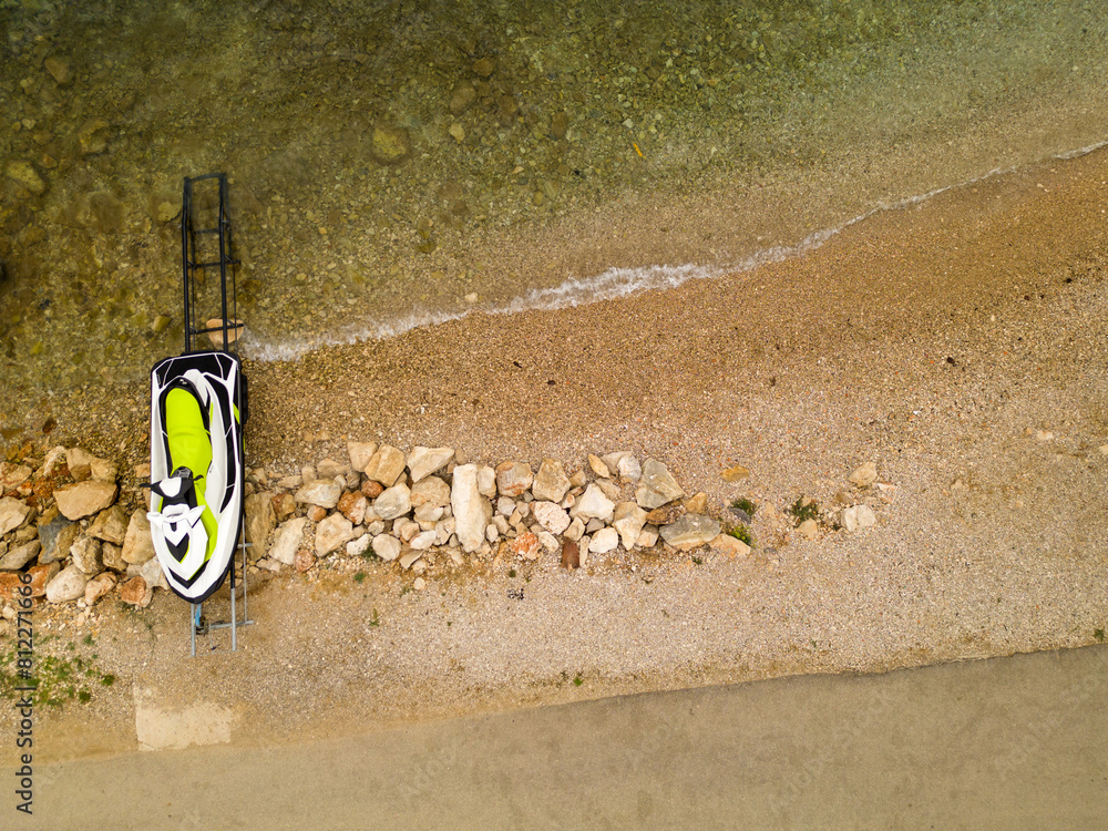 Obraz premium Aerial view of a jet ski parked on a rocky beach shoreline. The clear water and sandy beach create a perfect summer holiday vibe, ideal for adventure and water sports themes.