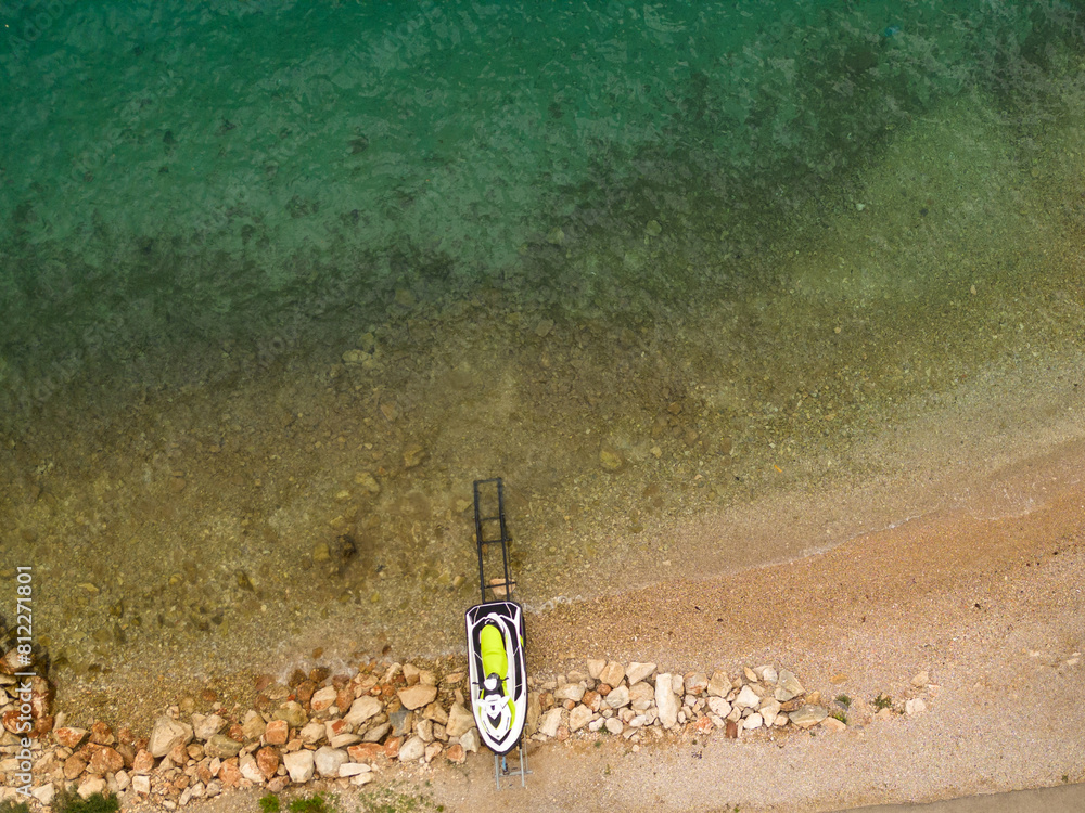 Aerial view of a jet ski parked on a rocky beach shoreline. The clear water and sandy beach create a perfect summer holiday vibe, ideal for adventure and water sports themes.