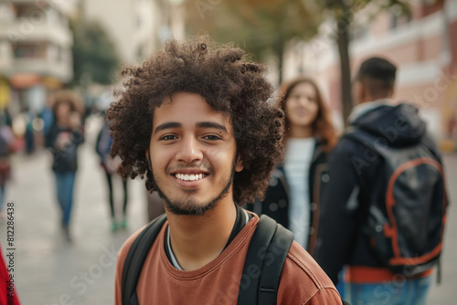 Cheerful young man with curly hair and a backpack smiling towards the camera, with blurry figures of diverse people walking behind him in an urban, lively street setting