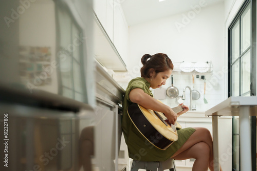 Focused Asian woman playing acoustic guitar in a modern kitchen, sitting on a stool