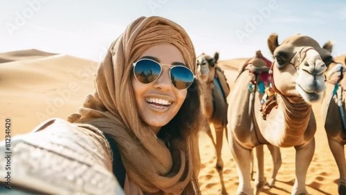 Young Moroccan woman embraces Sahara adventure taking selfies with camels in golden dunes. Concept Travel, Adventure, Culture, Photography, Desert Safari. photo