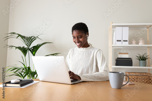 Black female entrepreneur working at home office. Happy African American woman using laptop and wireless earphones.