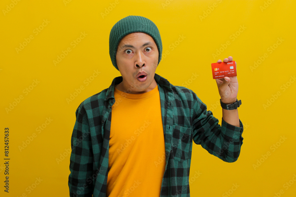 A surprised young Asian man, dressed in a beanie hat and casual shirt, holds a credit card while standing against a yellow background A surprised young Asian man, dressed in a beanie hat and casual shirt, holds a credit card while standing against a yellow background