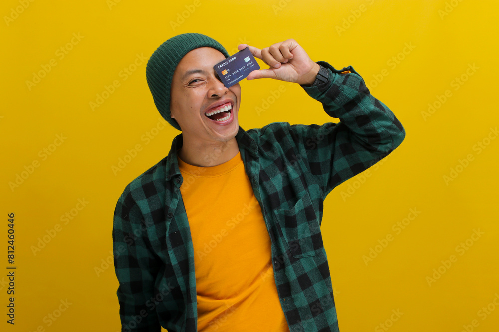 An excited young Asian man, wearing a beanie hat and casual shirt, covers one eye with a credit card, symbolizing concepts of purchase, consumerism, money, and payment, isolated on yellow background An excited young Asian man, wearing a beanie hat and casual shirt, covers one eye with a credit card, symbolizing concepts of purchase, consumerism, money, and payment, isolated on yellow background