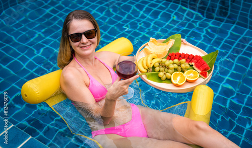 Beautiful woman lying on floating hammock in the swimming pool with wine and tray of fruits © romaset