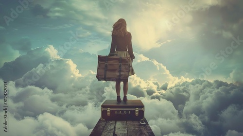 A woman stands on top of a vintage suitcase, gazing out at an expansive sky filled with clouds. Her back is to the camera, and she holds a large travel bag in her hand. She is dressed in a dark jacket photo