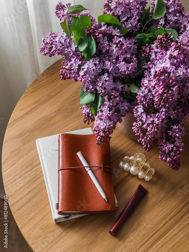 Women's table - notebooks, a hair clip, lip gloss and a bouquet of lilacs on a wooden table. Aesthetics lifestyle concept
