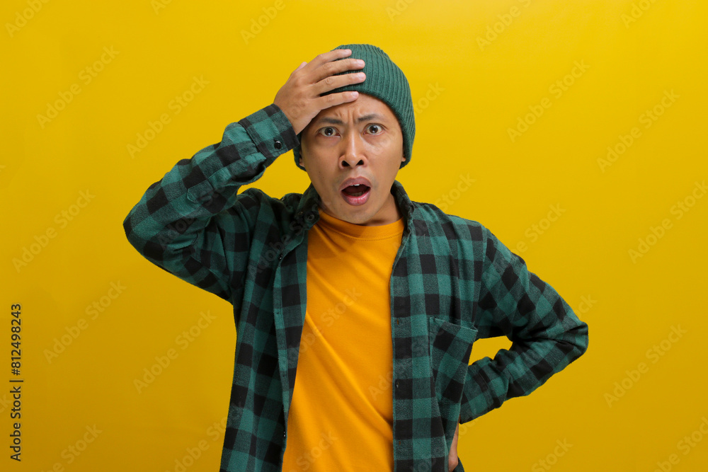Troubled young Asian man, dressed in a beanie hat and casual shirt, is depicted holding his hand to his forehead, appearing desperate and uncertain while standing against a yellow background Troubled young Asian man, dressed in a beanie hat and casual shirt, is depicted holding his hand to his forehead, appearing desperate and uncertain while standing against a yellow background