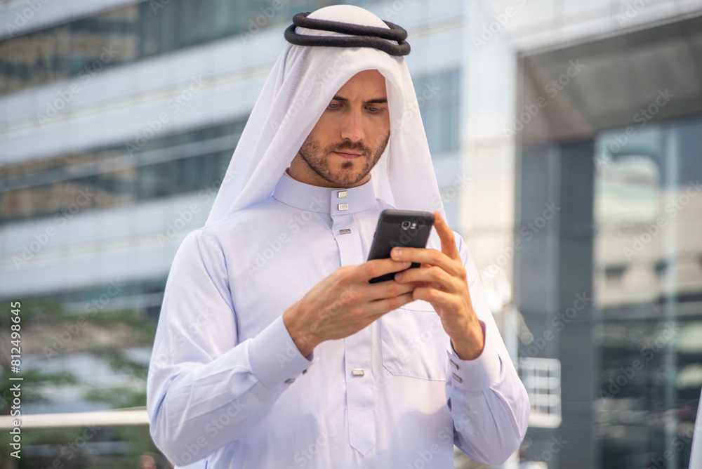 Arab male businessman wearing traditional colored clothes, holding a smartphone, typing a message, chatting on social media, standing outside the building.
