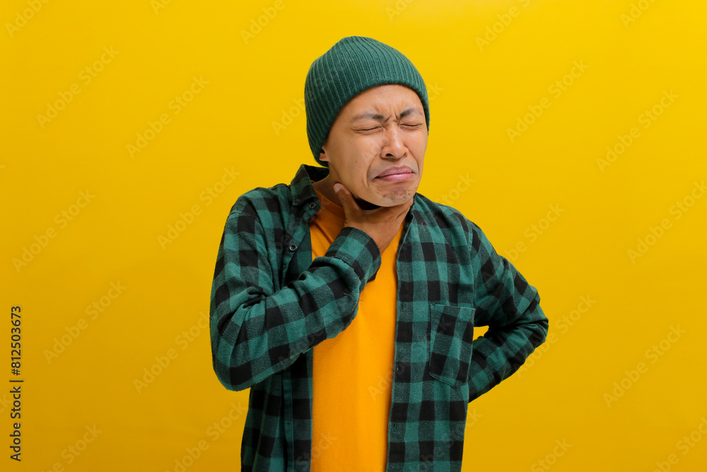 Young Asian man in beanie hat and casual shirt is experiencing a sore throat, possibly due to irritation or an infection, Isolated on yellow background. Healthcare and medical concept. Young Asian man in beanie hat and casual shirt is experiencing a sore throat, possibly due to irritation or an infection, Isolated on yellow background. Healthcare and medical concept.
