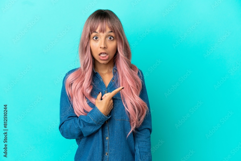 Young mixed race woman with pink hair isolated on blue background surprised and pointing side Young mixed race woman with pink hair isolated on blue background surprised and pointing side