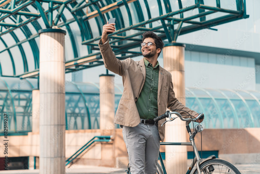 Cheerful happy handsome man with glasses taking selfie on the bike in front of business building. Cheerful happy handsome man with glasses taking selfie on the bike in front of business building.