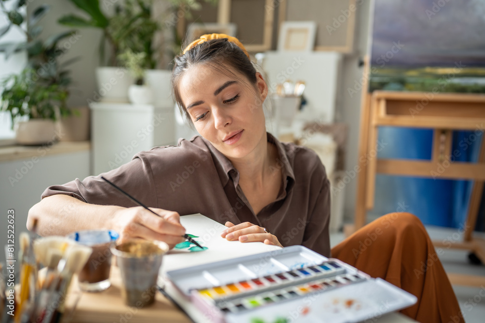 Fototapeta premium Calm female painter drawing with watercolor. Inspired pensive dreamy woman working on painting with focus immersed in creative process. Girl artist freely uses brushes, aquarelle to paint masterpieces