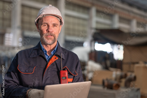 Confident senior male engineer wearing white hard hat and safety gloves smiles while working on heavy machinery in large workshop.