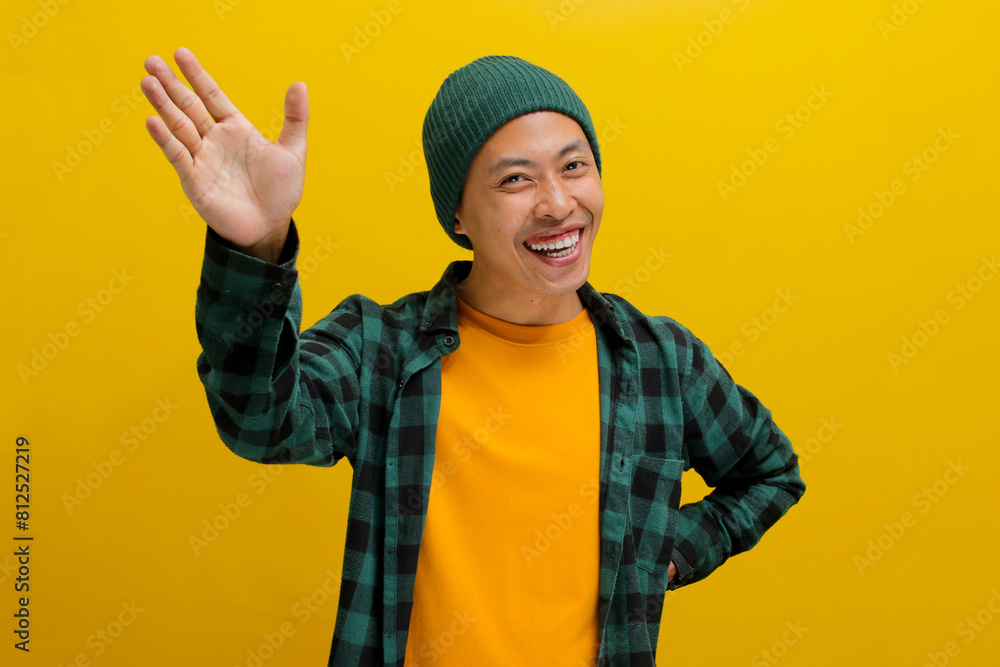 Young Asian man, dressed in a beanie hat and casual shirt, waves, saying hello with a happy and smiling face, offering a friendly welcome gesture while standing against a yellow background