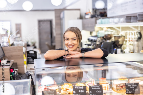 A cute female cafe owner is leaning against a glass display case with cakes and smiling.. Young female barista standing behind the bar in cafe smiling © weyo